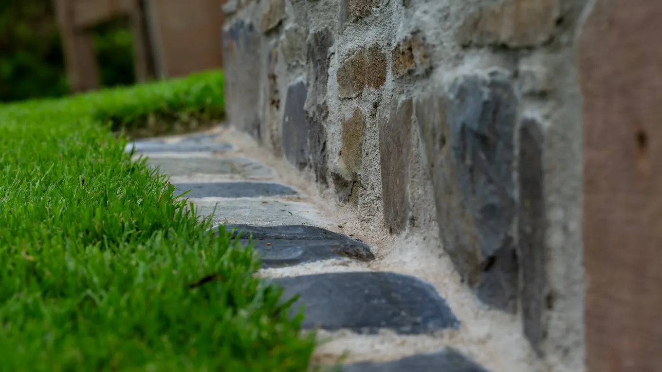Rasenkante aus dunklen Pflastersteinen entlang einer Natursteinmauer