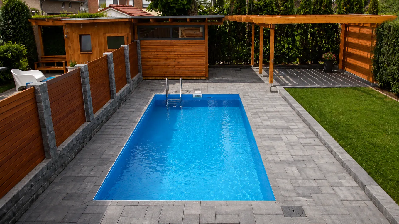 Rechteckiger Pool mit Edelstahlleiter in einer grauen Pflasterfläche, umgeben von Holzsichtschutz, Pergola und Gartenhaus