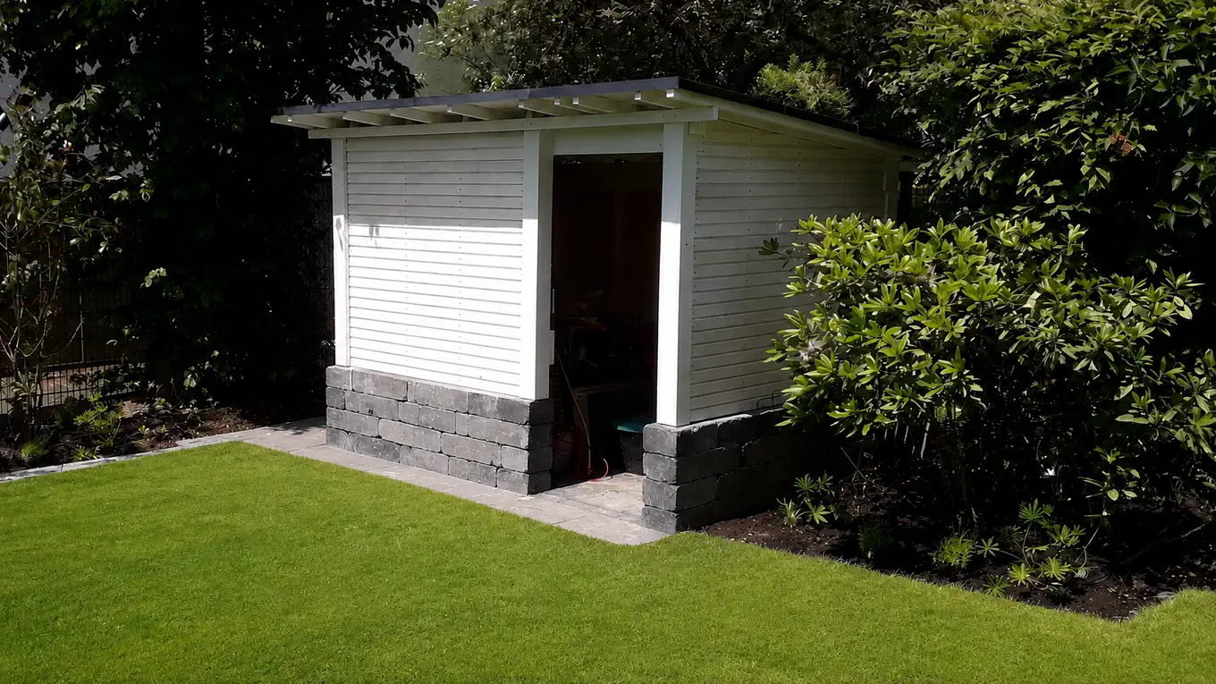 Weißes Gartenhaus mit Pultdach und offener Tür auf einem Sockel aus dunklem Mauerwerk, davor ein gepflasterter Streifen und eine Rasenfläche, rechts ein Rhododendron