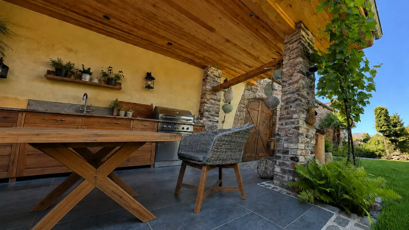 Überdachter Freisitz mit Holzdecke und Säule aus altem Feldbrandstein, Außenküche mit Spüle und Gasgrill, davor ein langer Holztisch mit gekreuztem Gestell und Korbsessel