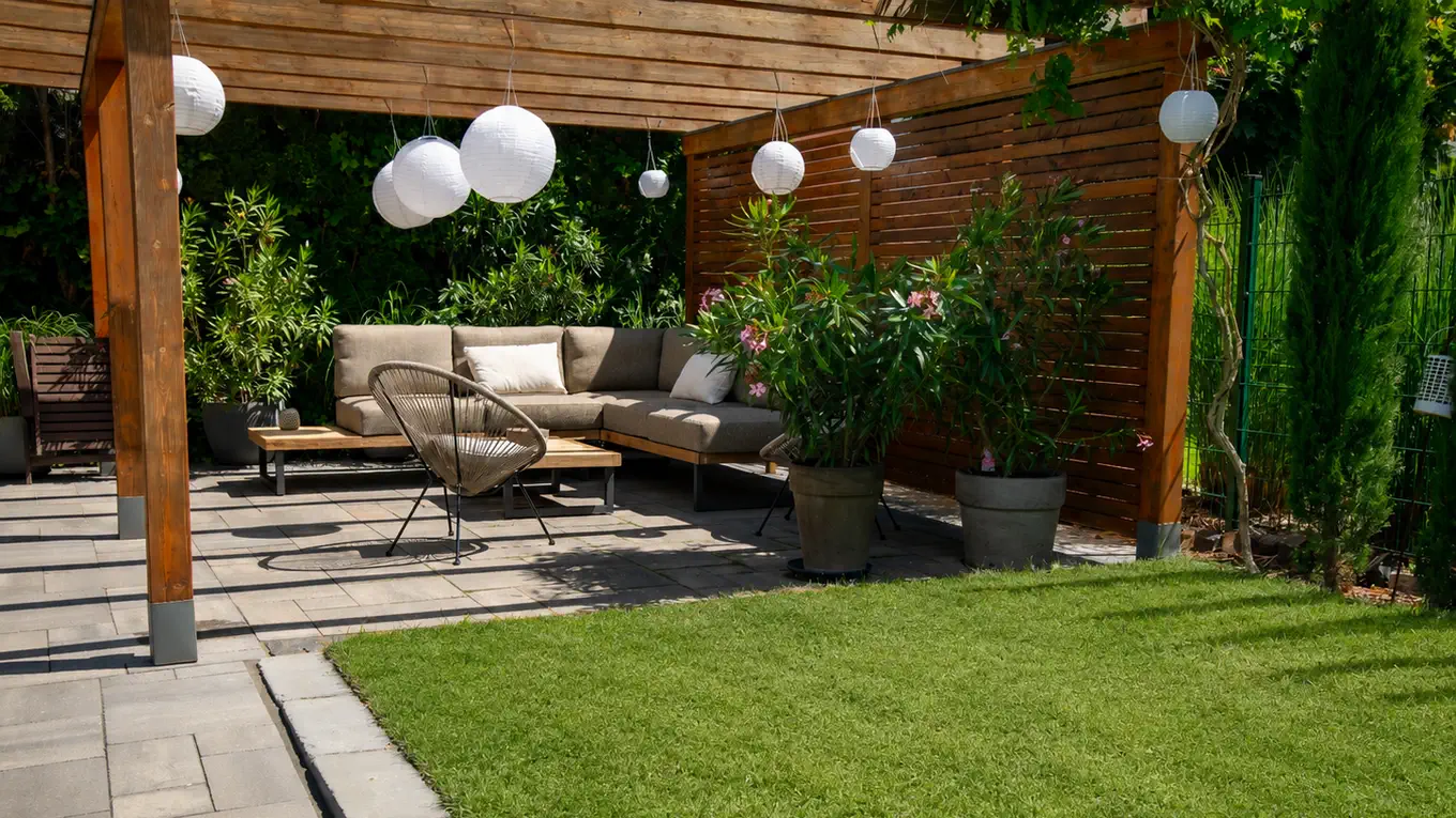 Sitzplatz unter einer Holzpergola mit Lampions und Sichtschutzwand, Loungesofa und Oleander in Kübeln, davor Plattenbelag mit sauberer Kante zum Rasen