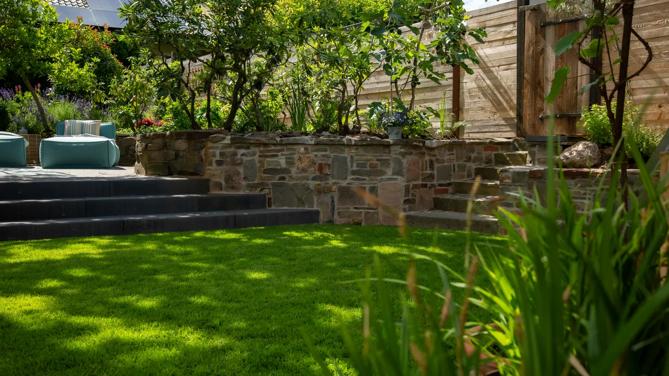 Terrassierter Garten mit Natursteinmauern, dunklen Blockstufen, Rasenfläche und Holzsichtschutz