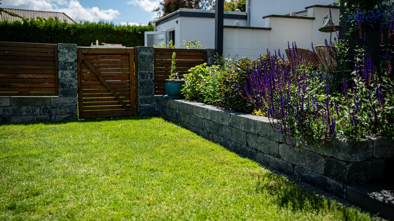 Holztor zwischen Natursteinsäulen in einer Gartenecke, davor Rasen und ein bepflanztes Hochbeet